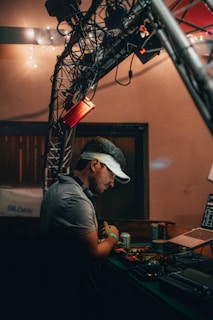 A person wearing a visor stands at a DJ booth, surrounded by equipment including a laptop and audio controls. The setting appears to be dimly lit, with string lights in the background and a truss structure overhead.