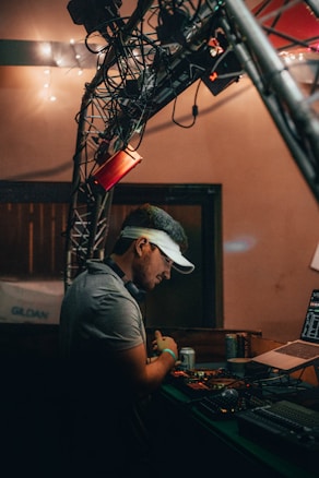 A person wearing a visor stands at a DJ booth, surrounded by equipment including a laptop and audio controls. The setting appears to be dimly lit, with string lights in the background and a truss structure overhead.