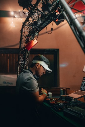 A person wearing a visor stands at a DJ booth, surrounded by equipment including a laptop and audio controls. The setting appears to be dimly lit, with string lights in the background and a truss structure overhead.