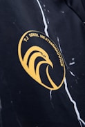 A close-up view of a sports logo on a fabric surface featuring an abstract eagle in a circular emblem. The logo includes the text 'TJ SOKOL HEJTMÁNKOVICE' in yellow against a dark background, with a pattern of white harlequin-like lines.
