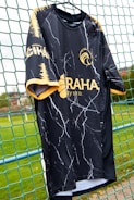 A black sports jersey with a marbled white pattern hangs on a net fence. The jersey features yellow accents on the sleeves and collar, and logos in yellow on the front and side. The background includes a blurred, grassy field.