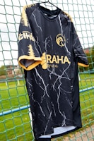 A black sports jersey with a marbled white pattern hangs on a net fence. The jersey features yellow accents on the sleeves and collar, and logos in yellow on the front and side. The background includes a blurred, grassy field.