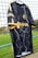 A black sports jersey with a marbled white pattern hangs on a net fence. The jersey features yellow accents on the sleeves and collar, and logos in yellow on the front and side. The background includes a blurred, grassy field.