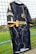 A black sports jersey with a marbled white pattern hangs on a net fence. The jersey features yellow accents on the sleeves and collar, and logos in yellow on the front and side. The background includes a blurred, grassy field.