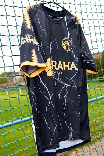 A black sports jersey with a marbled white pattern hangs on a net fence. The jersey features yellow accents on the sleeves and collar, and logos in yellow on the front and side. The background includes a blurred, grassy field.