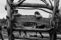 Two children sit on a rustic wooden structure, surrounded by a natural outdoor setting with trees in the background. They appear relaxed, with one wearing a plaid shirt and the other in a sweater, leaning casually on the wooden beam.