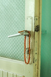 A white wooden door with a metal doorknob has a string of red-orange beads hanging from the handle. The door features a textured glass panel, and the overall setting has a vintage feel with muted colors.