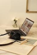 A cozy workspace features an open laptop displaying a grid of images, positioned on a light marble table. In the foreground, a black Sony camera rests on a stack of books, while a small vase of white flowers is slightly blurred in the background.