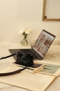A cozy workspace features an open laptop displaying a grid of images, positioned on a light marble table. In the foreground, a black Sony camera rests on a stack of books, while a small vase of white flowers is slightly blurred in the background.