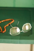 A pair of rectangular eyeglasses with thin metal frames lies on a green surface next to a string of orange beads. The glasses have a slight reflection on the lenses, and the beads appear to be made of a smooth, possibly amber-like material.