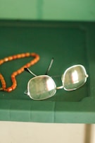 A pair of rectangular eyeglasses with thin metal frames lies on a green surface next to a string of orange beads. The glasses have a slight reflection on the lenses, and the beads appear to be made of a smooth, possibly amber-like material.