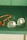 A pair of rectangular eyeglasses with thin metal frames lies on a green surface next to a string of orange beads. The glasses have a slight reflection on the lenses, and the beads appear to be made of a smooth, possibly amber-like material.