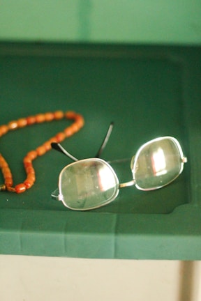 A pair of rectangular eyeglasses with thin metal frames lies on a green surface next to a string of orange beads. The glasses have a slight reflection on the lenses, and the beads appear to be made of a smooth, possibly amber-like material.