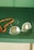 A pair of rectangular eyeglasses with thin metal frames lies on a green surface next to a string of orange beads. The glasses have a slight reflection on the lenses, and the beads appear to be made of a smooth, possibly amber-like material.