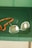 A pair of rectangular eyeglasses with thin metal frames lies on a green surface next to a string of orange beads. The glasses have a slight reflection on the lenses, and the beads appear to be made of a smooth, possibly amber-like material.