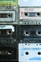 A collection of nine retro audio cassette tapes arranged in a grid pattern. The tapes feature diverse colors and labels, displaying a mix of transparent, black, and white plastic casings. Some tapes have handwriting on their labels, indicating personal collections or recorded music.