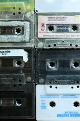 A collection of nine retro audio cassette tapes arranged in a grid pattern. The tapes feature diverse colors and labels, displaying a mix of transparent, black, and white plastic casings. Some tapes have handwriting on their labels, indicating personal collections or recorded music.