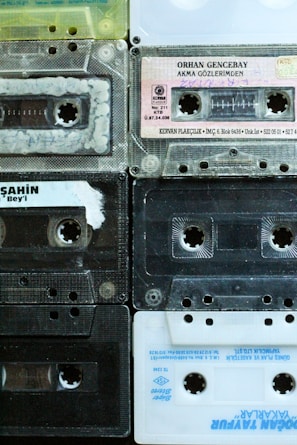 A collection of nine retro audio cassette tapes arranged in a grid pattern. The tapes feature diverse colors and labels, displaying a mix of transparent, black, and white plastic casings. Some tapes have handwriting on their labels, indicating personal collections or recorded music.