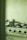 A kitchen with a gas stove featuring four burners, one of which is lit. A stainless steel pot with a lid is placed on the lit burner. The background shows a pale green tiled wall, and there are various kitchen items partially visible.