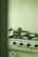 A kitchen with a gas stove featuring four burners, one of which is lit. A stainless steel pot with a lid is placed on the lit burner. The background shows a pale green tiled wall, and there are various kitchen items partially visible.