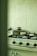 A kitchen with a gas stove featuring four burners, one of which is lit. A stainless steel pot with a lid is placed on the lit burner. The background shows a pale green tiled wall, and there are various kitchen items partially visible.