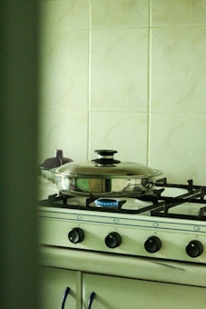 A kitchen with a gas stove featuring four burners, one of which is lit. A stainless steel pot with a lid is placed on the lit burner. The background shows a pale green tiled wall, and there are various kitchen items partially visible.