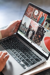 A person is operating a laptop, with the screen displaying a grid of photography images and graphics. The user interface appears to be for browsing or editing digital images. The laptop is placed on a wooden surface.