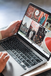 A person is operating a laptop, with the screen displaying a grid of photography images and graphics. The user interface appears to be for browsing or editing digital images. The laptop is placed on a wooden surface.