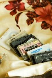 A collection of vintage cassette tapes is arranged on a cream-colored fabric with a floral pattern. The tapes are a mix of transparent, black, and white casings, and some have visible labels with text. The floral design features red and brown flowers.