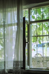 An open window with sheer curtains gently blowing. The view outside reveals green foliage and a parked vehicle partially visible. Intricate wrought iron bars are set within the window frame.