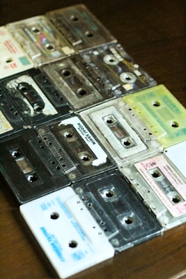 A collection of retro cassette tapes arranged in an overlapping pattern on a wooden surface. The tapes display a variety of colors and some have visible labels, indicating they have been used. The casings are mostly transparent or have solid colors such as black and white, with some showing signs of wear.