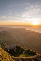 A breathtaking aerial view of a mountainous landscape at sunset, featuring a secluded building nestled among green hills. The sky is filled with warm, golden hues as the sun dips below the horizon, casting a serene glow over the expansive valley and distant body of water.
