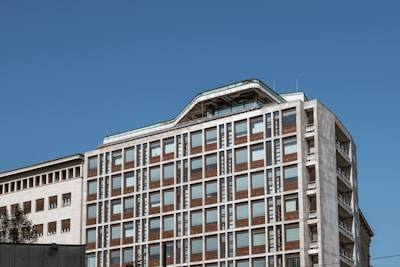 A multi-story building with a modern design featuring a grid-like pattern of windows. The architecture combines elements of glass and concrete, with a blue sky providing a backdrop. The building's top floors have an angled roof, adding to the architectural interest.