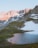 Rocky mountains with patches of snow are bathed in soft sunlight. A small, tranquil alpine lake reflects the warm hues of the surrounding landscape, bordered by green grassy areas.