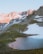 Rocky mountains with patches of snow are bathed in soft sunlight. A small, tranquil alpine lake reflects the warm hues of the surrounding landscape, bordered by green grassy areas.