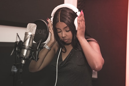 A person wearing headphones is standing in front of a large microphone. The setting appears to be a recording studio, with dark acoustic panels and professional audio equipment.