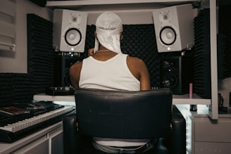 A person wearing a white tank top and head covering is seated in a home music studio. The setting includes two large white speakers, a keyboard, and soundproofing foam on the walls, creating a focused and creative environment.
