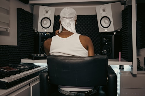A person wearing a white tank top and head covering is seated in a home music studio. The setting includes two large white speakers, a keyboard, and soundproofing foam on the walls, creating a focused and creative environment.