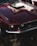 A classic, vintage car with a glossy, dark red finish is parked outdoors. The front view highlights the distinctive headlights, chrome bumper, and sleek lines that showcase its retro style.