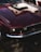 A classic, vintage car with a glossy, dark red finish is parked outdoors. The front view highlights the distinctive headlights, chrome bumper, and sleek lines that showcase its retro style.