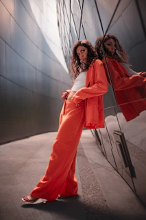 A woman with long, curly hair leans casually against a reflective metallic wall. She is wearing an orange pantsuit with a white top, and the wall creates a mirrored effect of her image. The setting is urban and modern, with smooth, sleek surfaces and soft lighting.