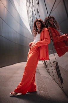 A woman with long, curly hair leans casually against a reflective metallic wall. She is wearing an orange pantsuit with a white top, and the wall creates a mirrored effect of her image. The setting is urban and modern, with smooth, sleek surfaces and soft lighting.