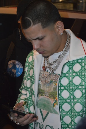 A man with a distinctive haircut and jewelry, including large chains and a pendant, is looking intently at a smartphone. He wears a patterned green and white jacket over a graphic t-shirt.