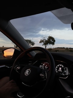 A car interior with a view of the dashboard and steering wheel. The dashboard displays illuminated dials and a central digital screen showing navigational information. Outside, a scenic landscape with silhouetted trees and a colorful sunset reflected in the side mirror is visible.