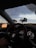 A car interior with a view of the dashboard and steering wheel. The dashboard displays illuminated dials and a central digital screen showing navigational information. Outside, a scenic landscape with silhouetted trees and a colorful sunset reflected in the side mirror is visible.