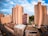 Tall residential buildings rise against a backdrop of a vibrant blue sky. The architecture features a mix of light-colored facades and red-painted sections. Roofs and balconies are visible, along with a few surrounding trees and a clear street below with parked cars.