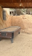 An old, weathered pool table sits outdoors on sandy ground, next to an earthen wall. The area is shaded by a wooden roof, and there is a small decorative metal window on a nearby wall. In the background, palm trees and a dilapidated structure can be seen.