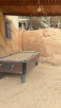 An old, weathered pool table sits outdoors on sandy ground, next to an earthen wall. The area is shaded by a wooden roof, and there is a small decorative metal window on a nearby wall. In the background, palm trees and a dilapidated structure can be seen.