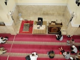 A group of people are seated on red and gold patterned prayer rugs in a large indoor space with cream-colored walls. A man is seated at the front behind a table with microphones, a book, and a cup, indicating he might be delivering a lecture or sermon. A wooden pulpit and a standing fan are nearby.
