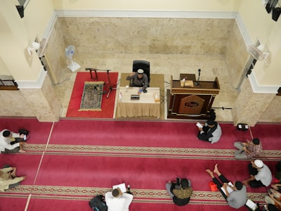 A group of people are seated on red and gold patterned prayer rugs in a large indoor space with cream-colored walls. A man is seated at the front behind a table with microphones, a book, and a cup, indicating he might be delivering a lecture or sermon. A wooden pulpit and a standing fan are nearby.